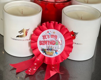以下が含まれることがあります： 「It's my birthday!」という文字と小さなケーキの絵が描かれた赤と白の誕生日ロゼット。ロゼットは、金のドラゴンロゴと「The Mystical Dragon」という文字が書かれた白いキャンドルのグループの前にある。