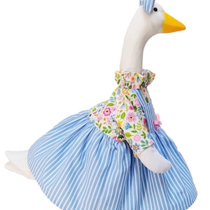 May include: A plush goose figurine wearing a blue and white striped dress with a floral print bodice and a matching blue and white striped bow on its head. The goose has a white body, a yellow beak, and black eyes, standing on a green base.