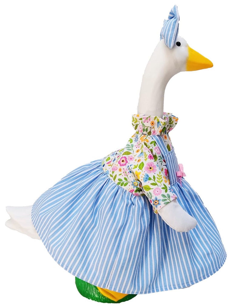 May include: A plush goose figurine wearing a blue and white striped dress with a floral print bodice and a matching blue and white striped bow on its head. The goose has a white body, a yellow beak, and black eyes, standing on a green base.