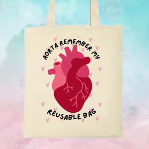 Può includere: Una borsa di tela beige con un cuore rosa e un'illustrazione di un cuore anatomico rosso. Il testo "AORTA REMEMBER MY REUSABLE BAG" è stampato sulla borsa.