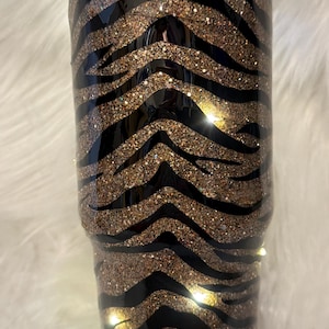 Könnte beinhalten: Ein zylindrischer Becher mit einem schwarz-goldenen Glitzer-Tigerstreifenmuster. Die Außenseite hat eine glatte, glänzende Oberfläche mit einem durchsichtigen Kunststoffdeckel. Das Design besteht aus abwechselnden horizontalen Streifen.