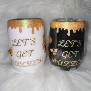 Könnte beinhalten: Zwei Edelstahlbecher mit goldenen Glitzer und einem tropfenden Honigdesign. Ein Becher ist weiß mit dem Text "Let's Get Buzzed" und der andere ist schwarz mit dem gleichen Text.