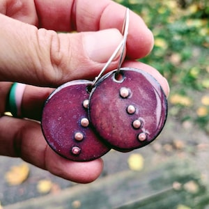 May include: A pair of dangle earrings with a dark purple ceramic disc and copper accents. The earrings are hanging from silver hooks.