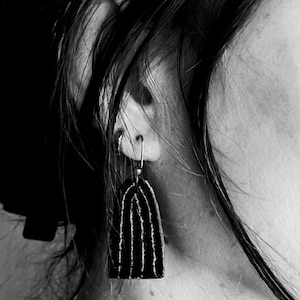 Può includere: Primo piano in bianco e nero di un orecchino. L'orecchino ha una forma scura e arcuata con linee bianche verticali. Pende da un cerchio d'argento. L'immagine mostra parte dell'orecchio e dei capelli di una persona.