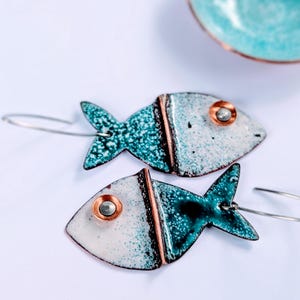 May include: A pair of handmade fish-shaped earrings. The earrings feature a teal and white speckled design with a copper accent. The fish have a small, round, copper-colored eye and a silver wire hook. A small teal bowl is in the background.