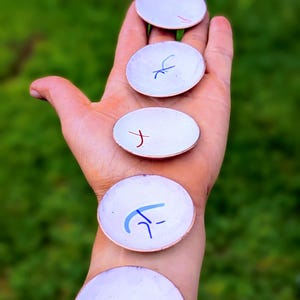 Pode incluir: Uma coleção de pequenos pratos redondos brancos com bordas de cobre. Cada prato apresenta um design abstrato único em vermelho, azul e preto. Os pratos são exibidos em um fundo verde desfocado.