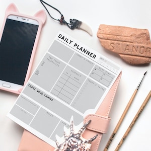 May include: A gray daily planner with sections for today's focus, schedule, meal plan, and three good things. The planner is open on a pink leather-bound notebook with a seashell on the bottom right corner. There are two paintbrushes and a small terracotta brick with the word "STANOR" on it.