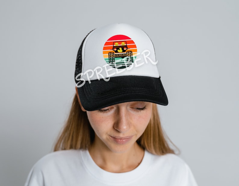 Może przedstawiać: Czarno-biała czapka trucker z białą siatką z tyłu. Z przodu czapki znajduje się kolorowa retro grafika zachodu słońca z sylwetką jeepa jadącego przez scenę.