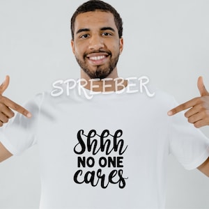 May include: A white t-shirt with black text that reads "Shhh no one cares". A person is wearing the shirt and pointing to it with both hands.
