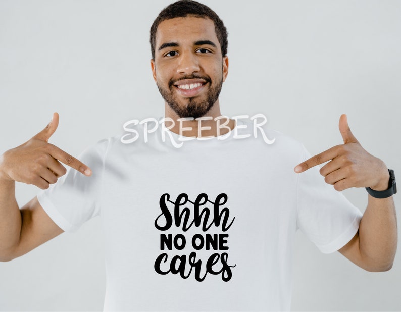 May include: A white t-shirt with black text that reads "Shhh no one cares". A person is wearing the shirt and pointing to it with both hands.