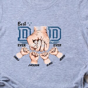 May include: Gray t-shirt with a blue graphic of a fist with the word "DAD" in blue letters and the words "Best Ever" on either side.  The fist is surrounded by four smaller fists with the names "Emma", "Jackson", "Mark", and "Henry" written below.