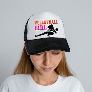 May include: A black and white trucker hat with a white front panel. The front panel features a silhouette of a volleyball player with the text "VOLLEYBALL GIRL SPREADER" in orange and pink.