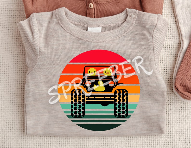 Może przedstawiać: Szary t-shirt z grafiką w stylu retro przedstawiającą jeepa z dwoma ż&oacute;łtymi gumowymi kaczkami w środku, jadącego przez zach&oacute;d słońca.