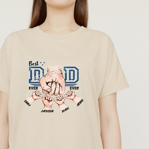 May include: A beige t-shirt with a blue and white graphic that says "Best Dad Ever" with four hands forming a fist around the word "Dad". The hands have names written below them: Emma, Jackson, Mark, and Henry.