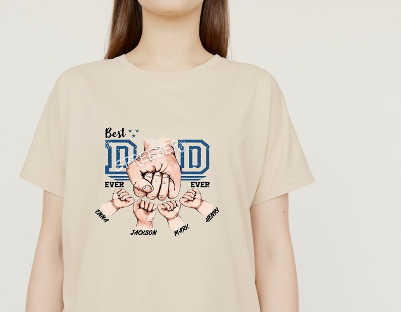 May include: A beige t-shirt with a blue and white graphic that says "Best Dad Ever" with four hands forming a fist around the word "Dad". The hands have names written below them: Emma, Jackson, Mark, and Henry.