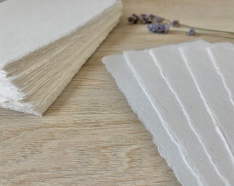 May include: A stack of white handmade paper sheets with deckled edges. The paper is arranged in a neat pile on a light wood surface.