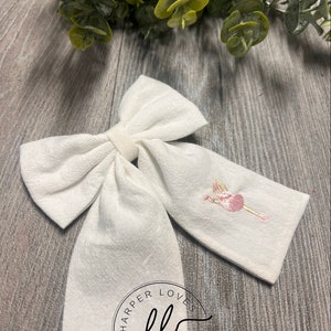 May include: White fabric bow with a pink ballerina embroidered on one side. The bow has a circle logo with the text "Harper Loves" and the initials "hl" in the center.