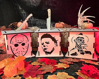 May include: Three wooden Halloween-themed candle holders with different horror movie characters. The first holder features a hockey mask, the second features Michael Myers, and the third features Freddy Krueger. Each holder has a knife or weapon on top.