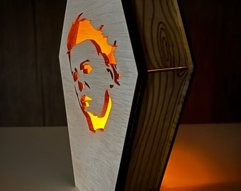 May include: A wooden coffin-shaped lamp with a cutout of a horror movie character's face. The lamp is lit from inside, casting a warm glow.