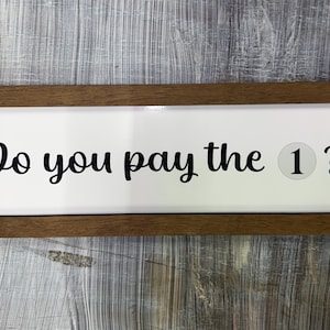 May include: A rectangular wooden framed sign with the text "Do you pay the 1 ?" in black script on a white background. The number 1 is in a circle. The sign is on a wooden surface.