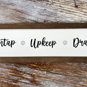 May include: A rectangular wooden sign with a white center and a dark brown frame. The sign displays the words "Untap", "Upkeep", and "Draw" in a black, handwritten font, with a die symbol between each word. A decorative item for tabletop gaming.