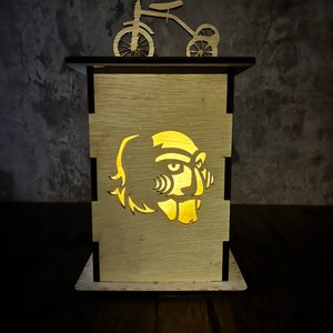May include: A wooden lamp with a cutout of a monster face. The lamp is lit and casting a warm glow. A wooden toy tricycle sits on top of the lamp.