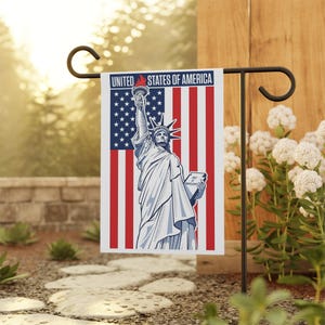 Puede incluir: Una bandera de jardín con fondo blanco y un diseño de bandera estadounidense roja, blanca y azul. La Estatua de la Libertad está en el centro de la bandera, sosteniendo una antorcha y un libro. El texto "UNITED STATES OF AMERICA" está impreso en la parte superior de la bandera.