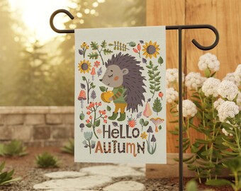 May include: A garden flag with a white background featuring a cartoon hedgehog holding an apple. The hedgehog is surrounded by colorful flowers, mushrooms, and leaves. The text "Hello Autumn" is printed in black at the bottom of the flag.