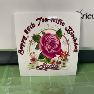 Peut inclure: Une carte blanche avec une rose rose et des accents dorés. Le texte "Happy 85th Tea-rrific Birthday Lydia" est écrit en rouge et noir.
