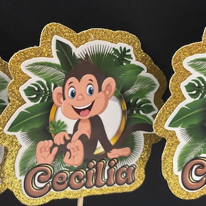 May include: Three gold glitter cake toppers with a jungle theme. Each topper features a cartoon monkey with brown fur, a green and brown leafy background, and a gold circle around the monkey. The toppers are personalized with the name "Cecilia".