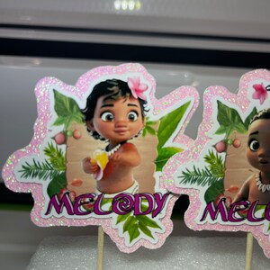 May include: Two pink glitter cupcake toppers featuring a cartoon baby girl with brown skin and dark hair. The baby is holding a yellow fruit. The toppers have green leaves and the name "Melody" written on them.