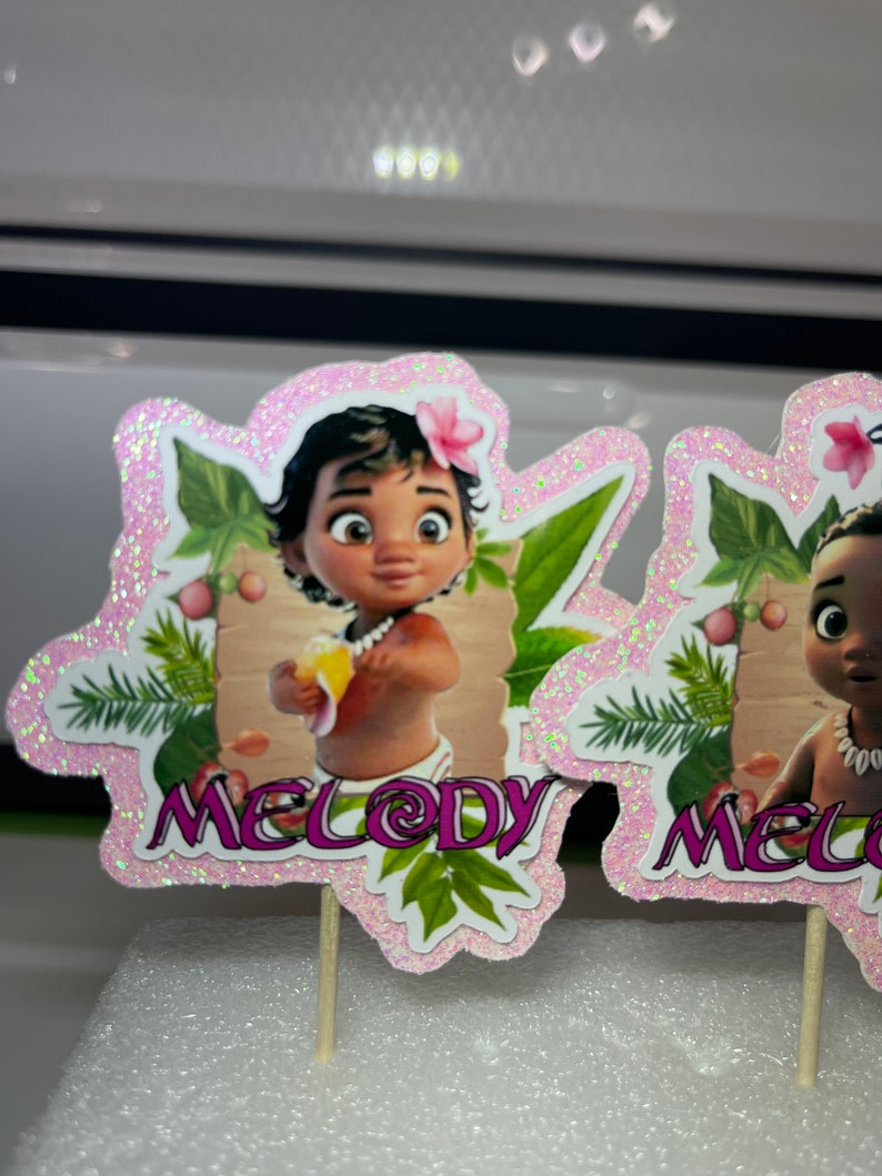 May include: Two pink glitter cupcake toppers featuring a cartoon baby girl with brown skin and dark hair. The baby is holding a yellow fruit. The toppers have green leaves and the name "Melody" written on them.