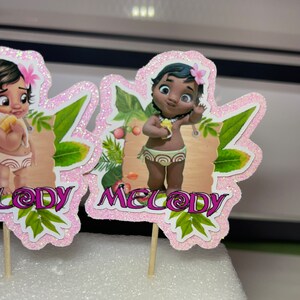 May include: Two pink and green cupcake toppers featuring cartoon baby girls with brown hair and brown eyes. The toppers are labelled "Lody" and "Melody".