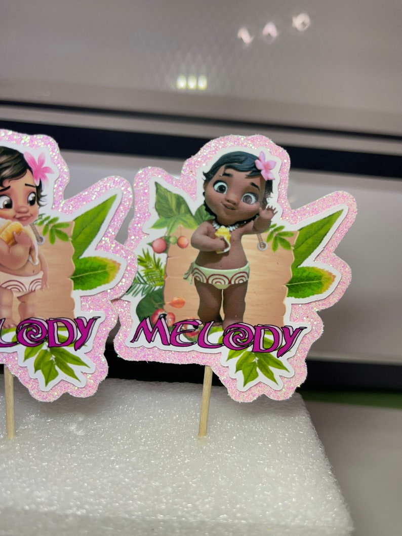 May include: Two pink and green cupcake toppers featuring cartoon baby girls with brown hair and brown eyes. The toppers are labelled "Lody" and "Melody".
