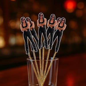 May include: A set of five clear, gold-glittered drink stirrers, each topped with a die-cut image of a person wearing a black tank top, black pants, and sunglasses, with arms crossed, standing in a clear glass tumbler on a wooden surface.