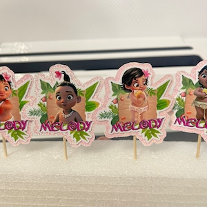 May include: Four pink and green cupcake toppers featuring a cartoon character with brown hair and brown eyes. Each topper has the word "Melody" printed on it.