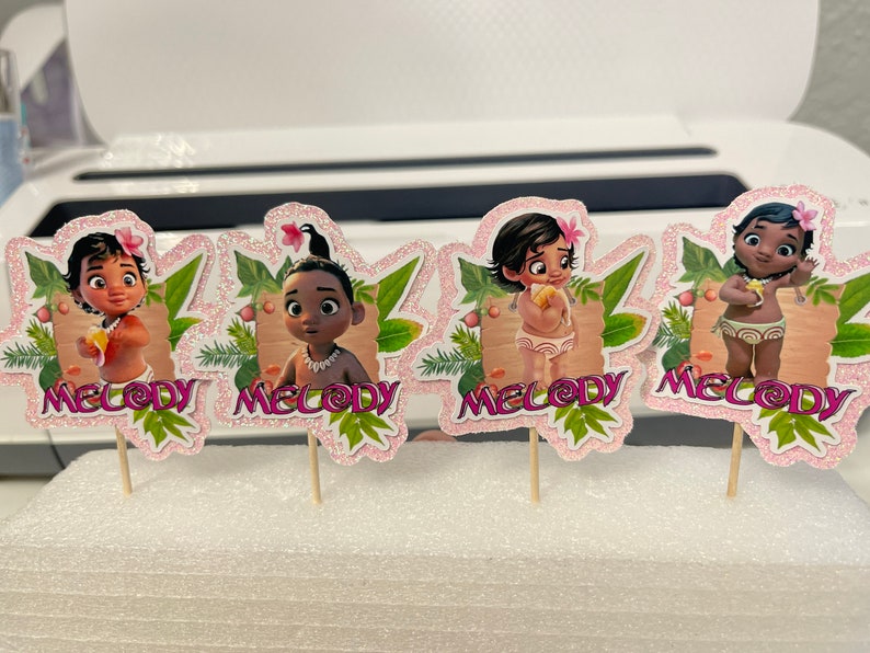 May include: Four pink and green cupcake toppers featuring a cartoon character with brown hair and brown eyes. Each topper has the word "Melody" printed on it.