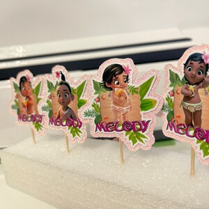 May include: Four cupcake toppers featuring a cartoon character with brown skin and black hair. Each topper has a pink background with glitter and green leaves. The character's name, "Melody", is written in pink letters on each topper.