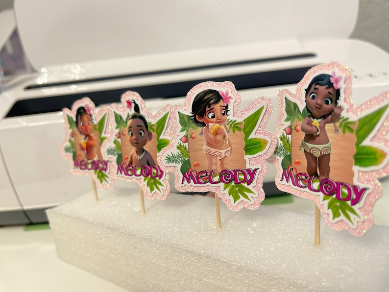May include: Four cupcake toppers featuring a cartoon character with brown skin and black hair. Each topper has a pink background with glitter and green leaves. The character's name, "Melody", is written in pink letters on each topper.