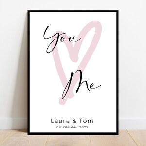 May include: A framed print with the words "You & Me" in elegant script over a light pink heart shape. Below, the names "Laura & Tom" and the date "08. Oktober 2022" are printed in black.