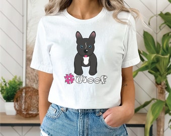 Op de afbeelding: Wit T-shirt met een cartoon illustratie van een zwart-grijze hond met blauwe ogen en een roze tong. De tekst "#Woof" staat onder de hond. Het shirt is gemaakt van een zacht materiaal.