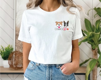 Op de afbeelding: Wit T-shirt met een kleine afbeelding van twee cartoonhonden en het woord "Best" in zwarte letters. De letter "s" is roze. De honden zijn een bruine en witte pitbull en een zwart-witte Boston Terrier.