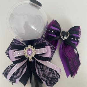 May include: A black and clear light stick with a large clear orb, adorned with black and lavender bows, lace, and a jeweled centerpiece. A matching bow with a heart-shaped jewel is also present.