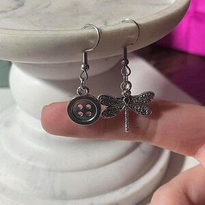 May include: Silver-coloured dangle earrings. One earring has a button design with four holes, and the other features a detailed dragonfly. The earrings are suspended from curved ear wires.