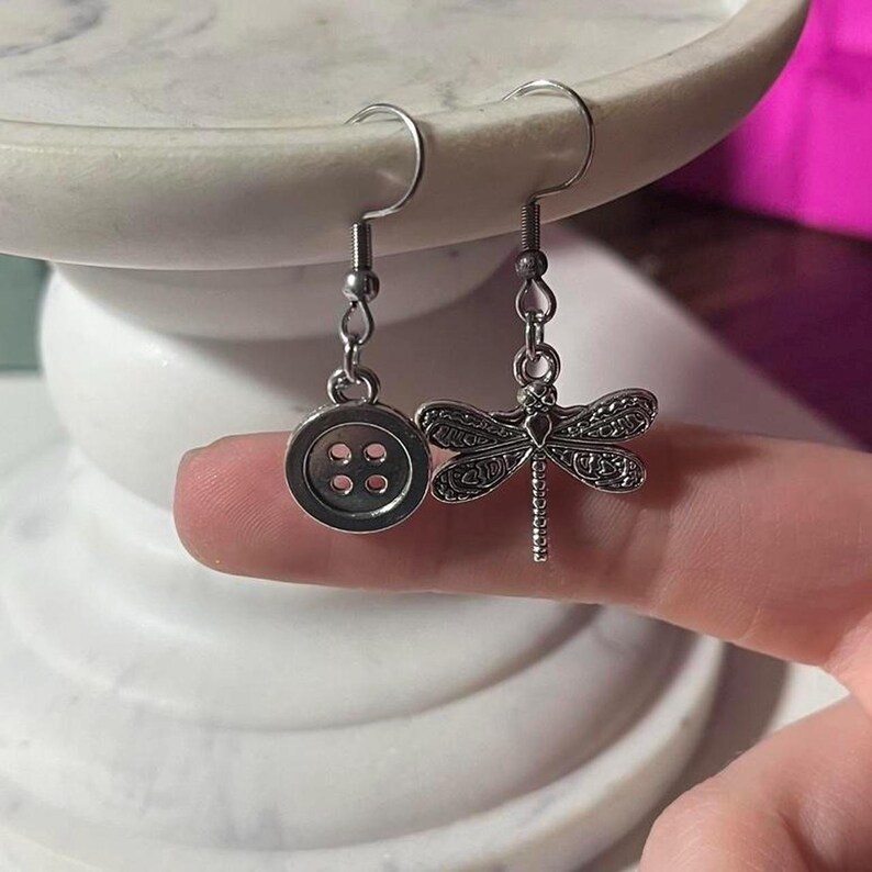 May include: Silver-coloured dangle earrings. One earring has a button design with four holes, and the other features a detailed dragonfly. The earrings are suspended from curved ear wires.