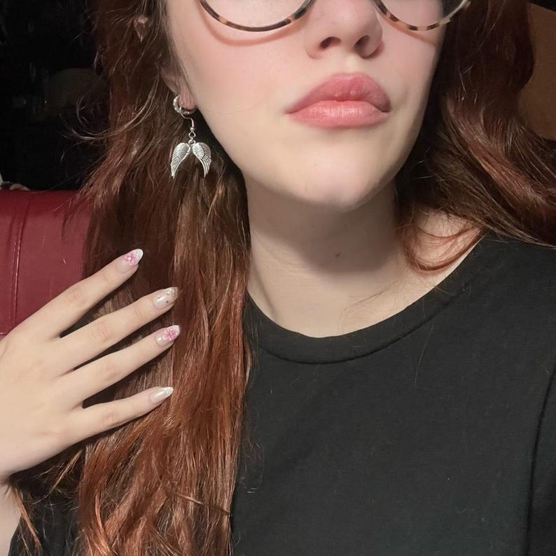 May include: Silver-toned angel wing earrings. Each earring features a detailed wing design, suspended from a small hoop. The earrings are shown against a blurred background, with a black top.