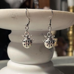 May include: A pair of silver-toned ladybug earrings. Each earring features a detailed ladybug charm with small, round cutouts and a crown-like design on top. The earrings hang from simple, curved ear wires.