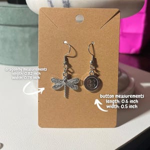 May include: Silver dragonfly and button earrings on a brown card. The dragonfly earring is 2.08 cm long and 1.98 cm wide. The button earring is 1.52 cm long and 1.27 cm wide. The earrings hang from silver-coloured hooks.