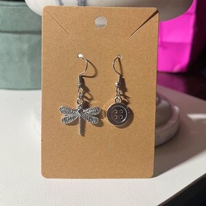 May include: A pair of silver-coloured dangle earrings. One earring features a detailed dragonfly charm, while the other has a button design with four small circles. The earrings are displayed on a brown card.