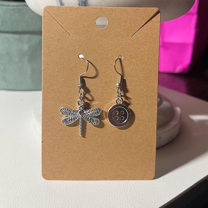 May include: A pair of silver-coloured dangle earrings. One earring features a detailed dragonfly charm, while the other has a button design with four small circles. The earrings are displayed on a brown card.